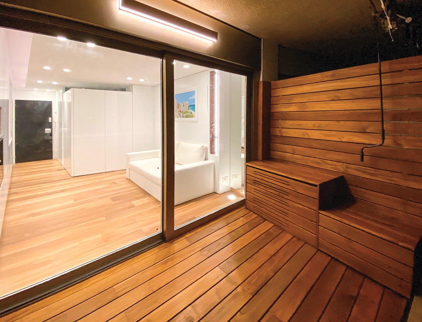 Terrazza con parquet in legno con porta scorrevole in vetro che conduce a una stanza dalle pareti bianche con divano. 