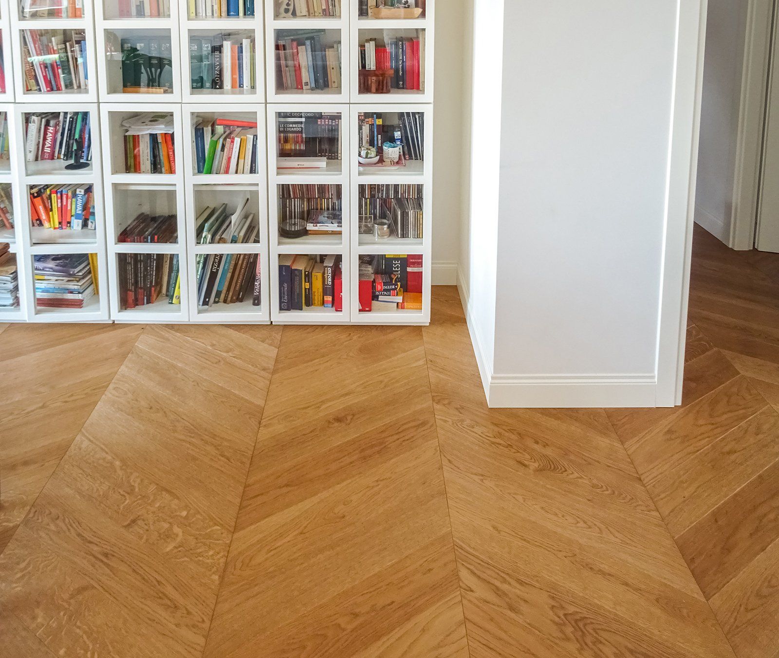 Pavimento in parquet con motivo a chevron, accanto a una libreria bianca e a una parete con una porta aperta.