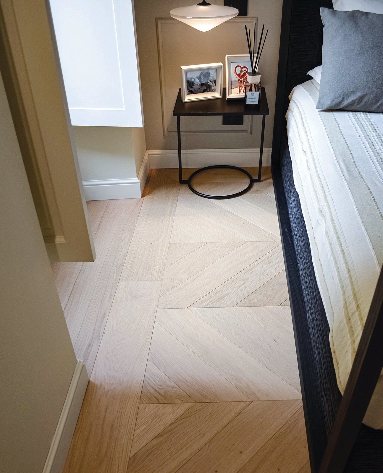 Pavimento in legno chiaro con motivo a zig-zag, accanto a un letto e un comodino decorati.