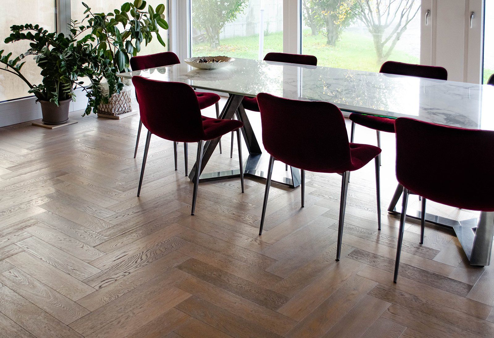 Sala da pranzo con tavolo in marmo, sedie bordeaux e pavimenti in legno. Ampia finestra, piante nell'angolo.