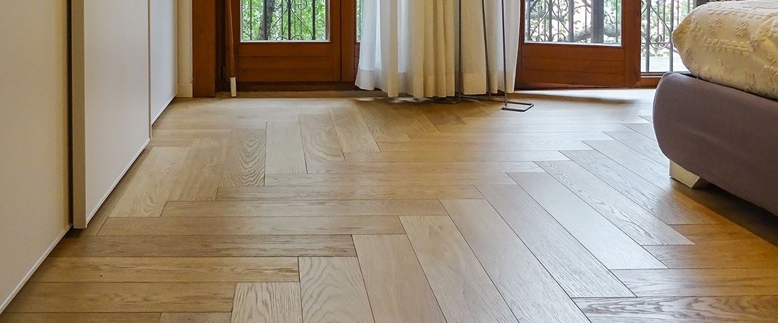 Pavimento in legno a spina di pesce in una stanza, con un letto visibile sulla destra e una porta e delle tende sullo sfondo.
