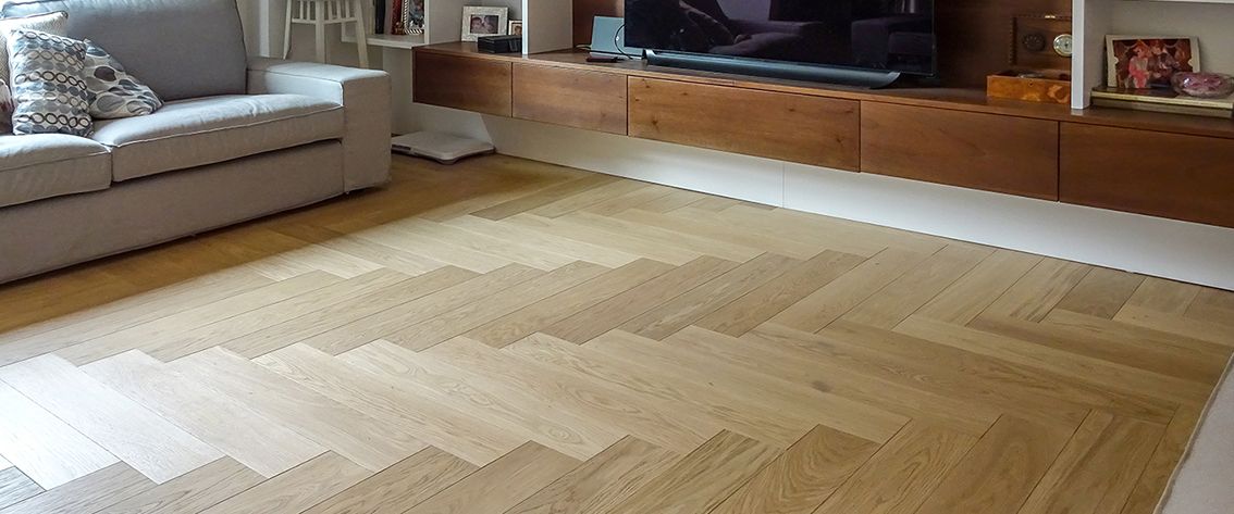 Pavimento in legno con motivo a chevron nel soggiorno, divano beige e mobile TV integrato.