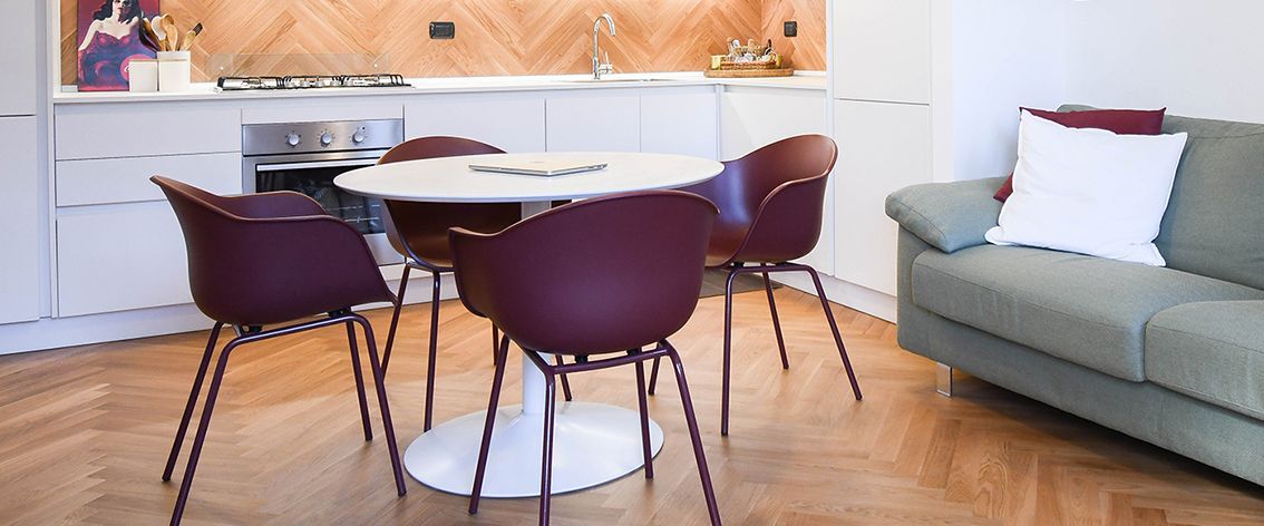 Una zona pranzo con un tavolo rotondo, quattro sedie bordeaux, un divano verde e una cucina con pavimento in legno.