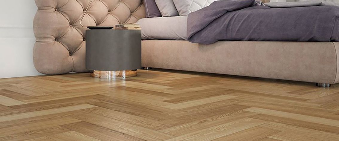 Pavimento in legno a spina di pesce davanti a un letto con comodino grigio e testiera trapuntata.