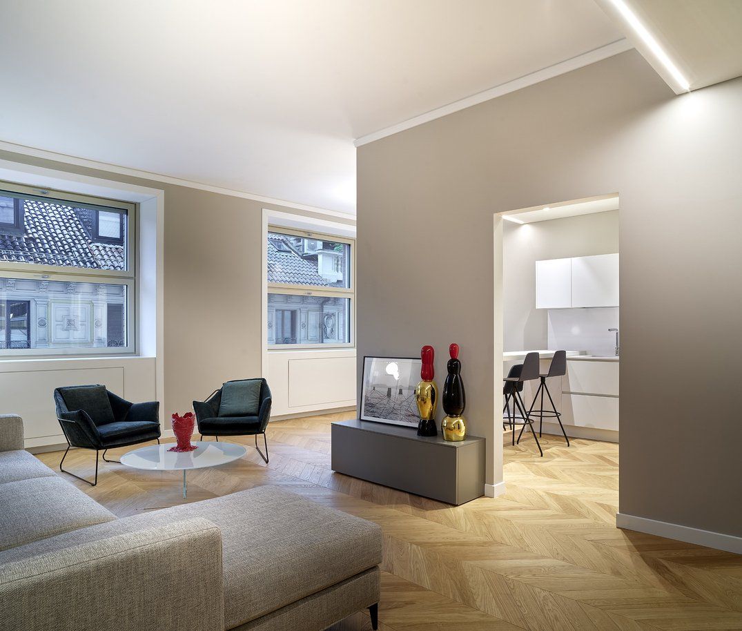 Soggiorno moderno con pareti beige, pavimenti a spina di pesce, divano grigio, poltrone e cucina visibile sulla porta.