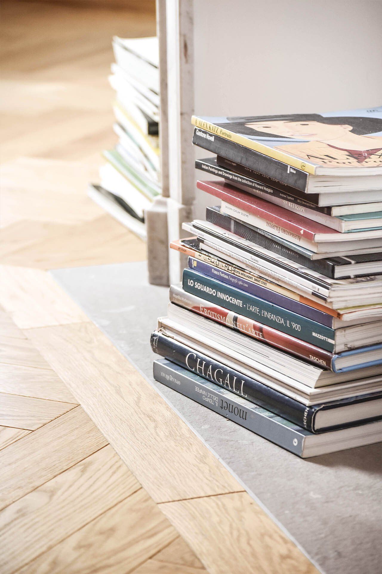 Pila di libri e riviste accanto a una porta su un pavimento in legno con motivo a spina di pesce.