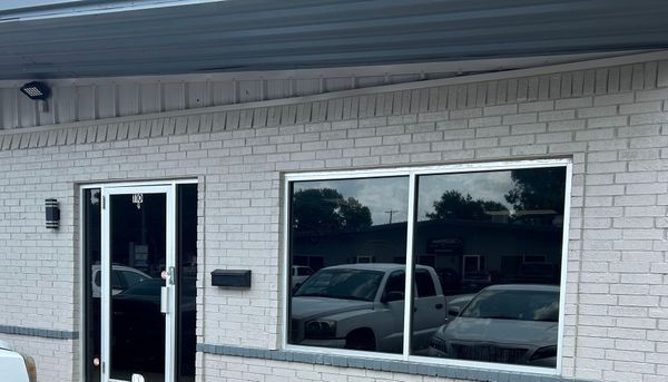 White brick storefront with glass door and large windows reflecting parked cars under an awning