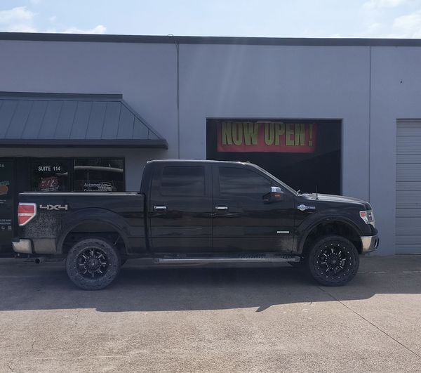 Black lifted pickup truck parked outside a gray shop with a “Now Open” sign.
