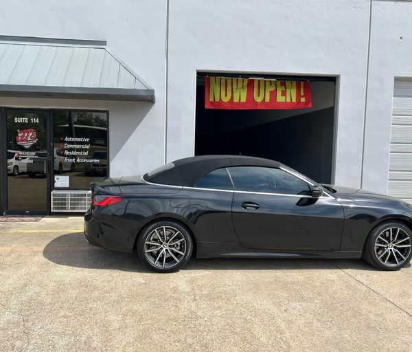 Black sports car parked outside a storefront with a bright “NOW OPEN!” sign.