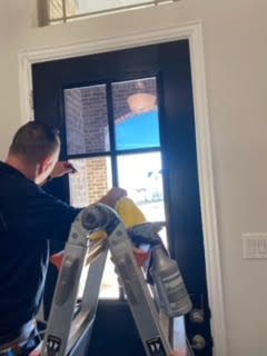 Person on a ladder installing or adjusting a black front door with glass panes indoors.