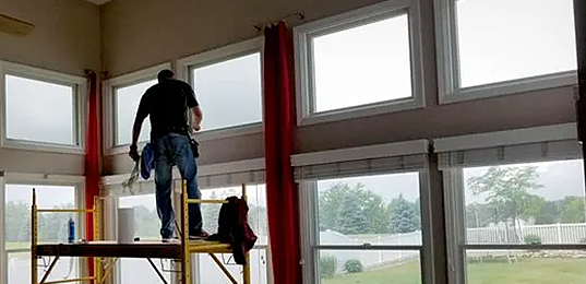 Worker standing on a scaffold inside a room with large windows, painting or installing trim near the ceiling