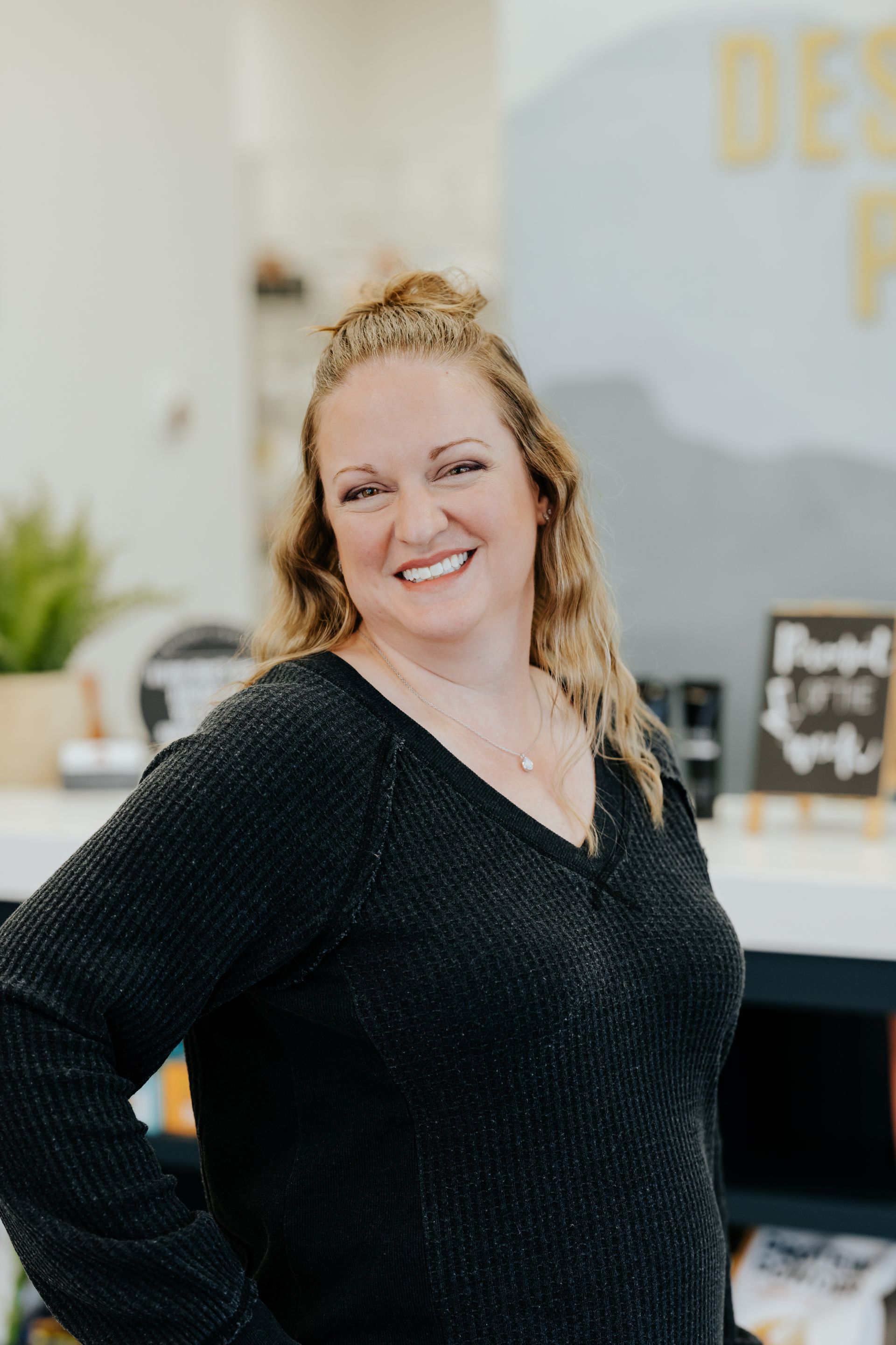 Woman with blonde hair smiling, wearing a black sweater, in a store with decor.