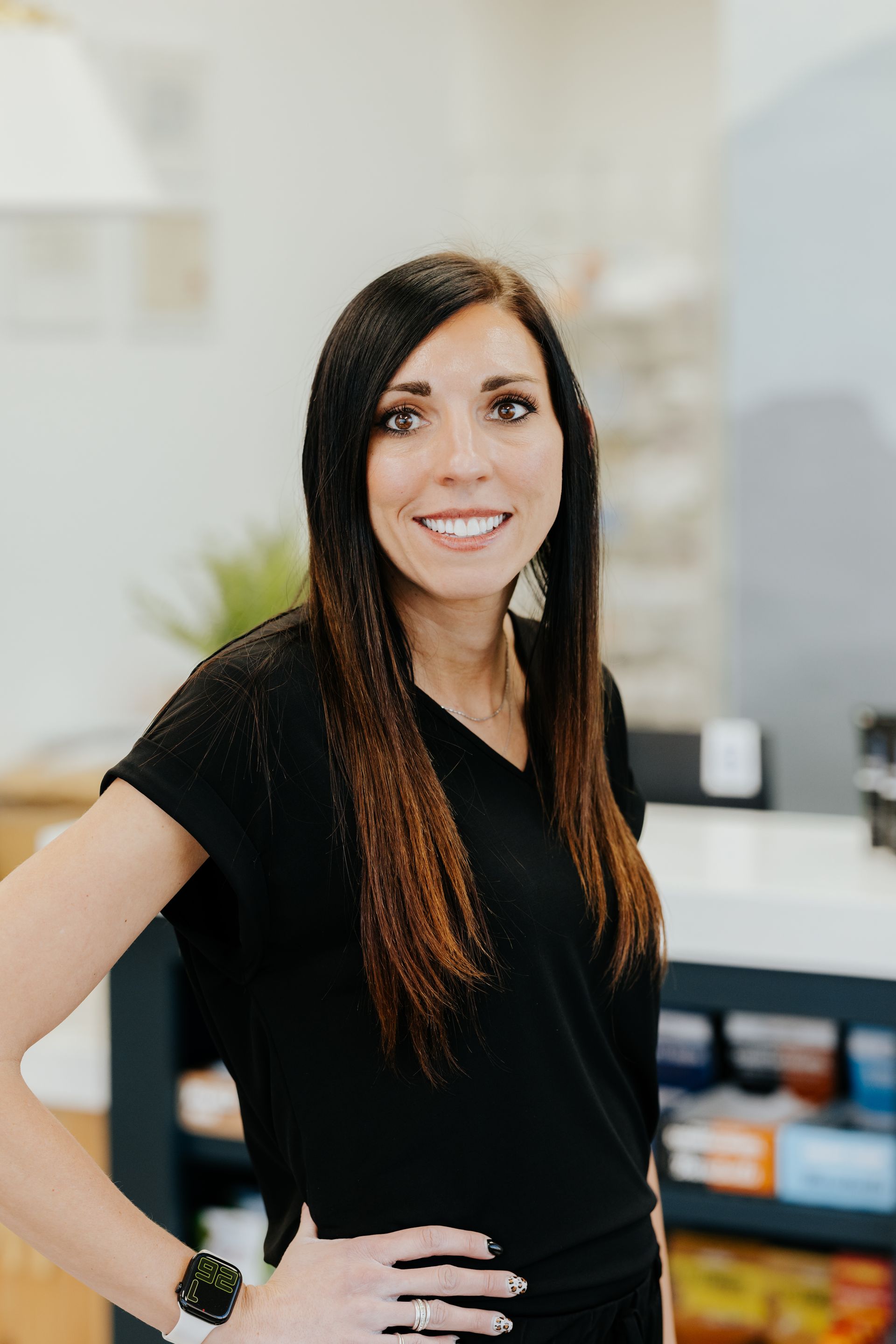 Woman with long brown hair, smiling, wearing a black top, hand on hip, in a store.