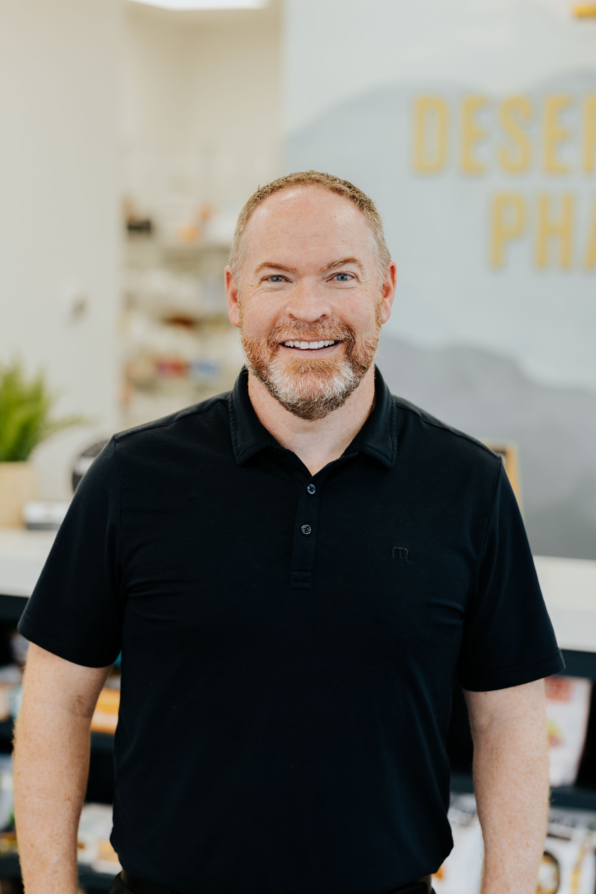 Man with a beard in a black polo shirt smiles in a brightly lit setting; background has yellow text and shelves.
