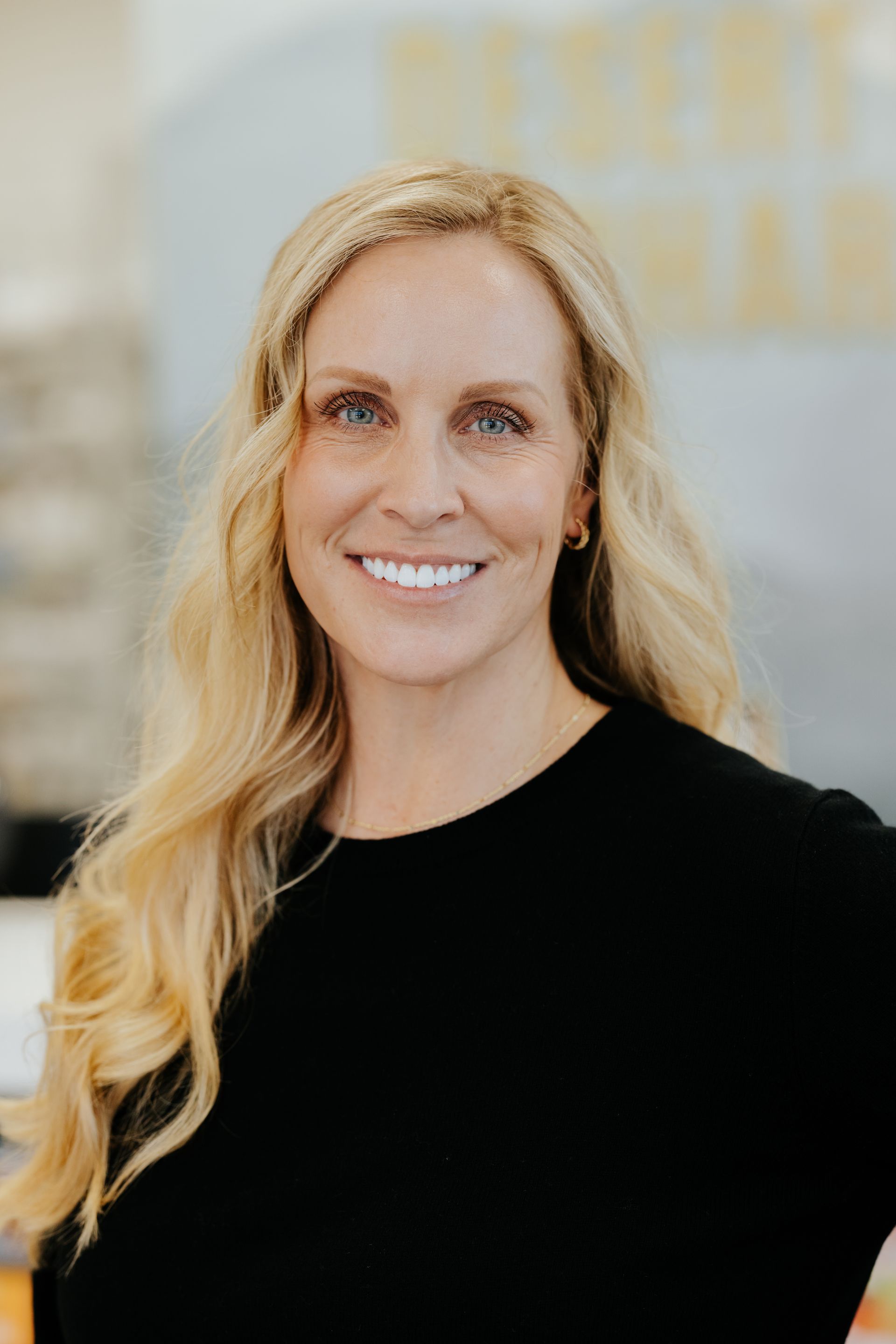 Woman with blonde hair smiling, wearing a black shirt, in a brightly lit room.
