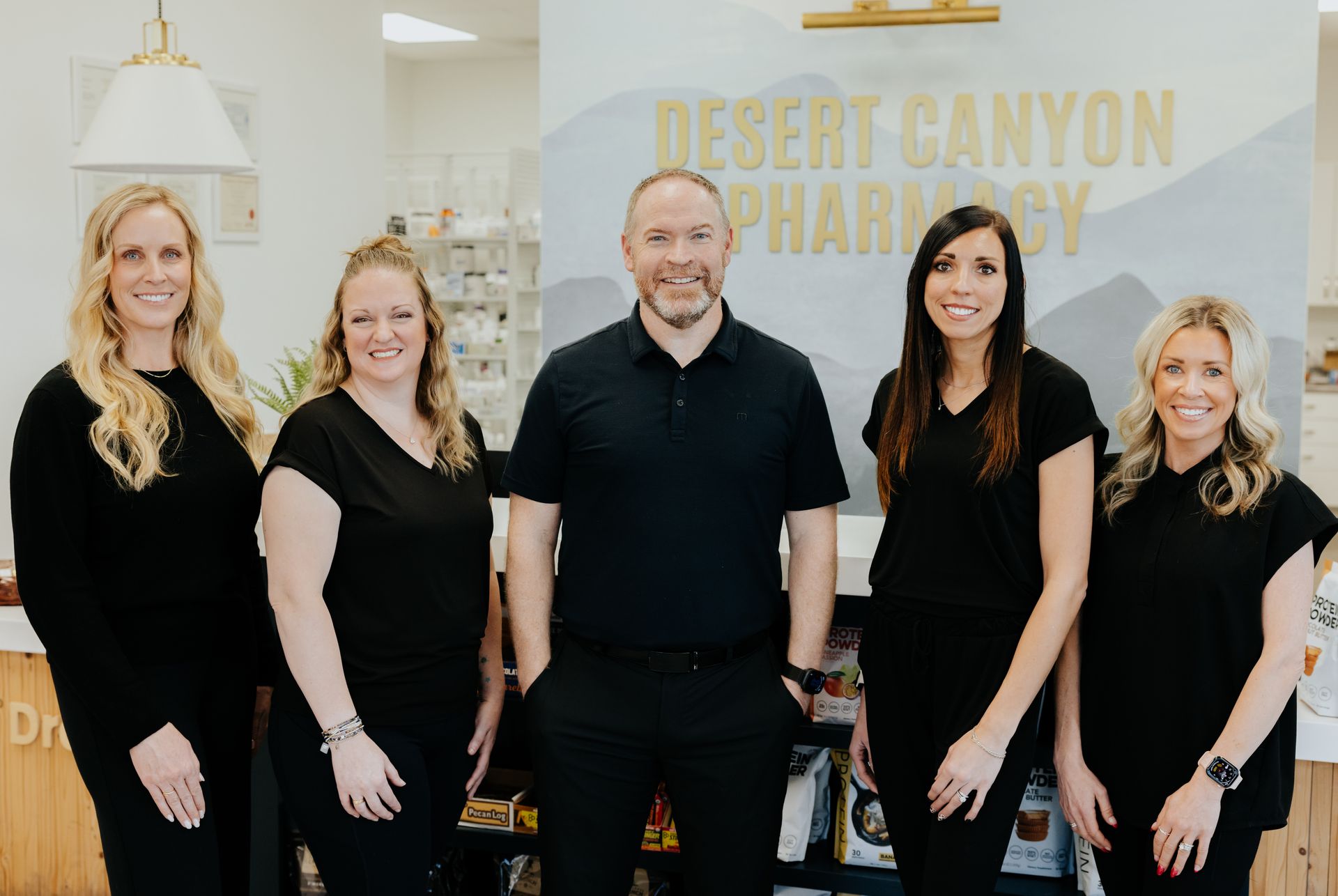 Five pharmacy employees in black shirts pose in front of a counter and sign reading 