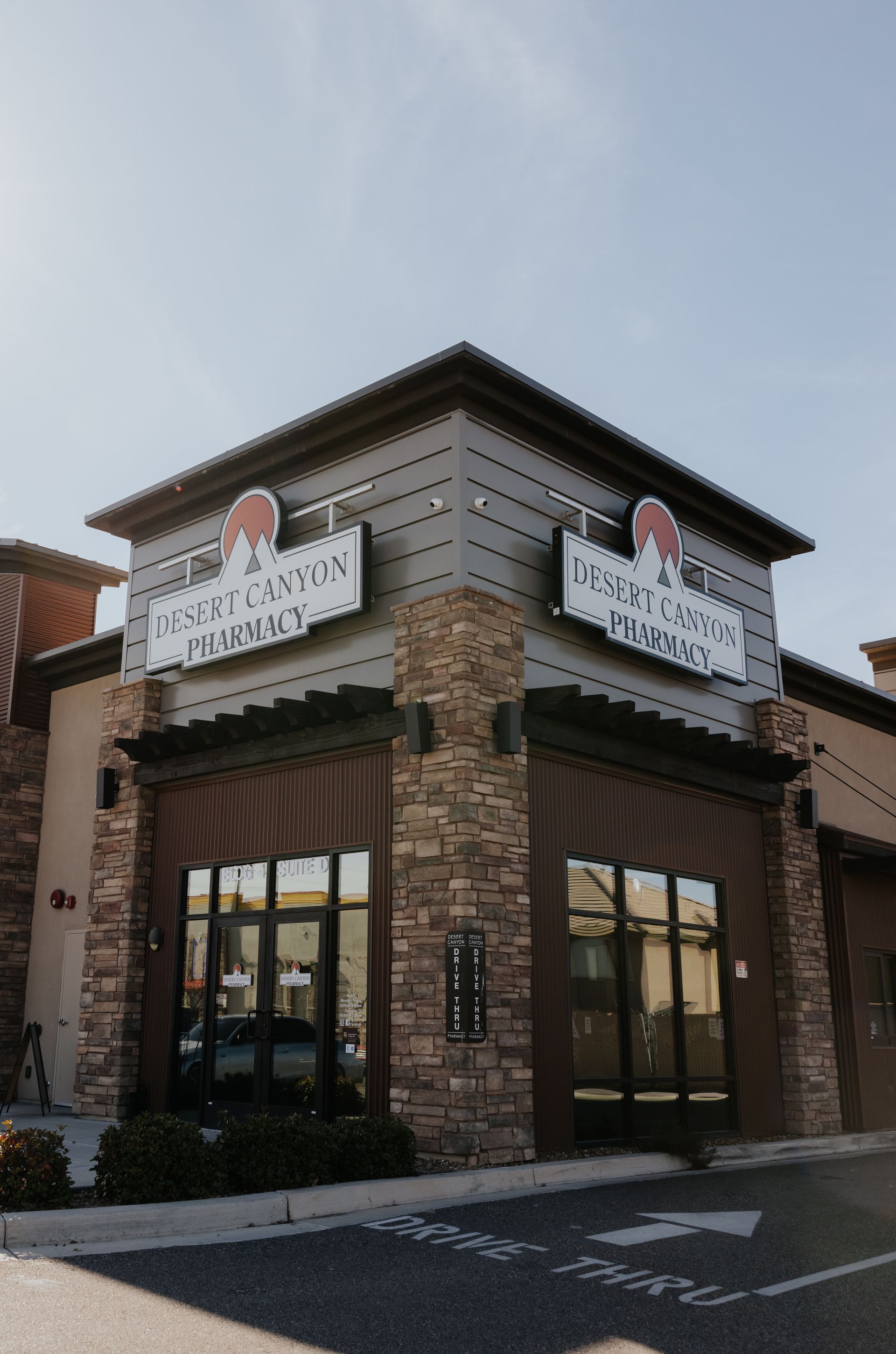 Exterior of a pharmacy building with signage. Brown brick and gray siding. Sunlight.