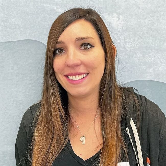 Woman with long brown hair smiles, wearing a black shirt with a white-striped zipper, and a necklace. Light blue background.