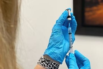 Gloved hands filling a syringe from a vial, against a white wall and a framed picture.