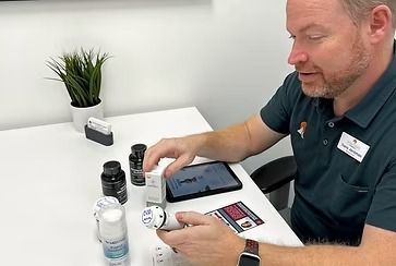 A man in a green shirt demonstrating products on a white table. Includes bottles, boxes, and a tablet.