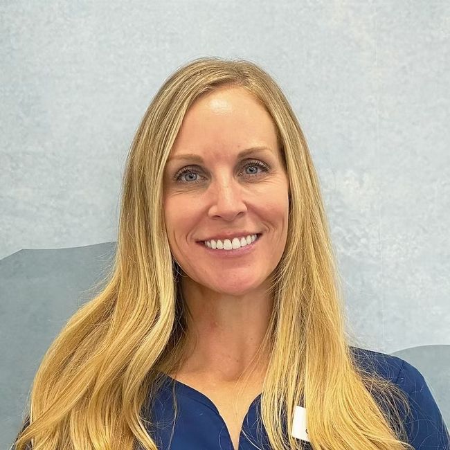 Woman with blonde hair smiling, wearing a blue top, against a blue and gray wall.