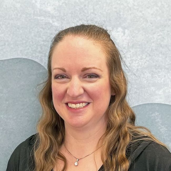 Woman with long wavy hair smiling in front of a blue and white patterned wall. She wears a black top.