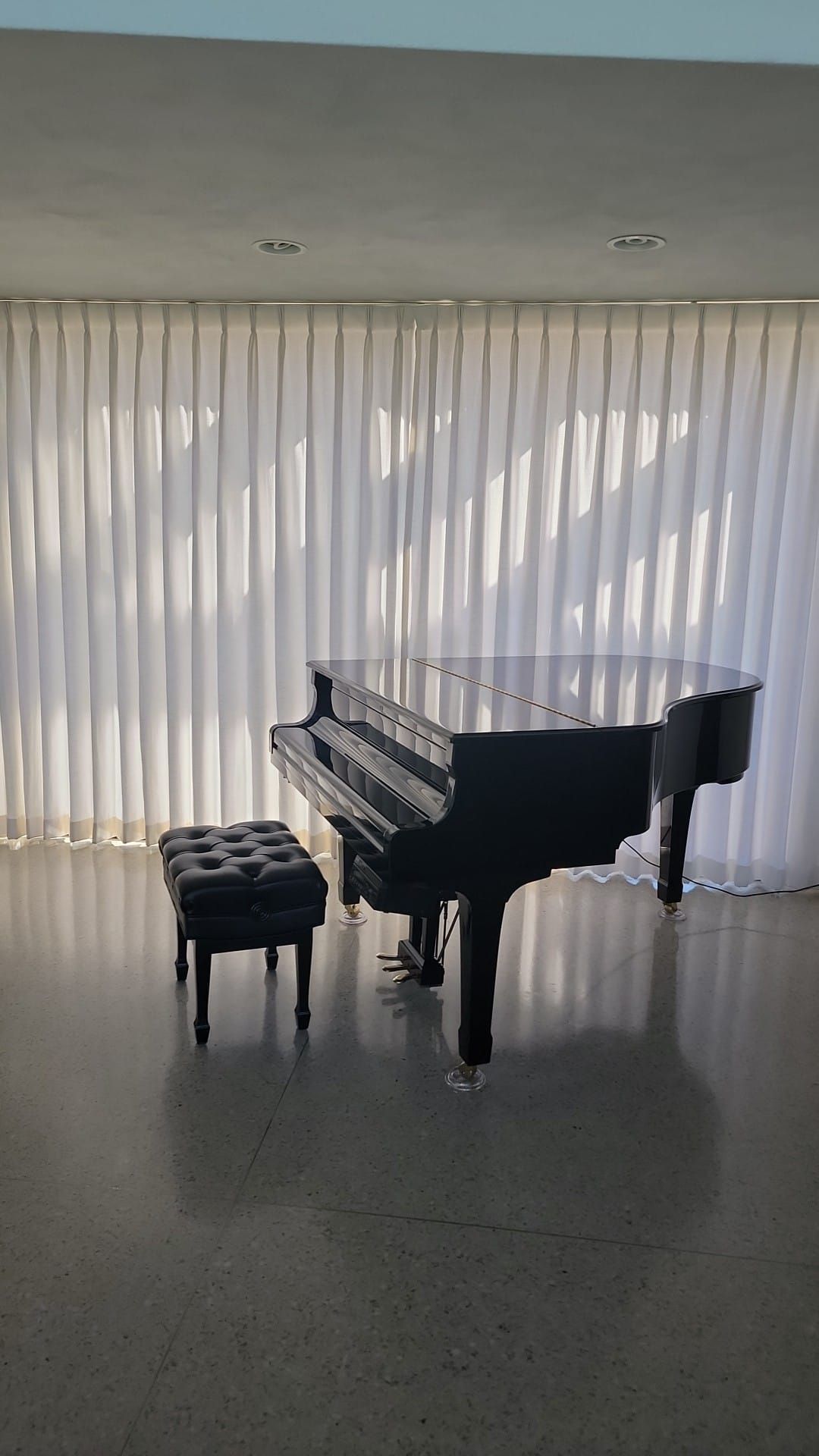 A black grand piano is sitting in front of a window with white curtains.