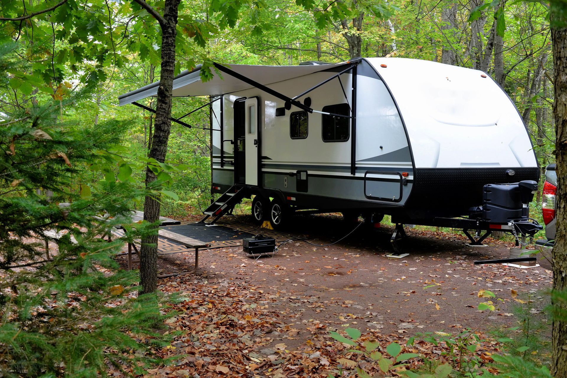 Caravane blanche avec auvent garée dans un camping boisé, parmi les arbres verts et les feuilles mortes