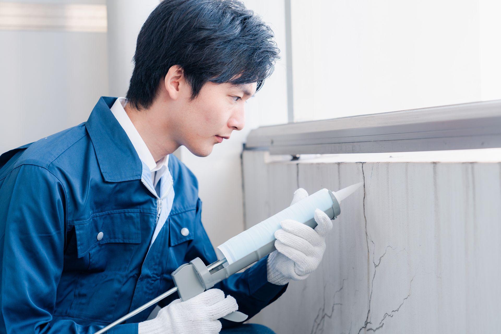 Un ouvrier en combinaison bleue applique du mastic sur un mur fissuré à l'aide d'un pistolet à calfeutrer.