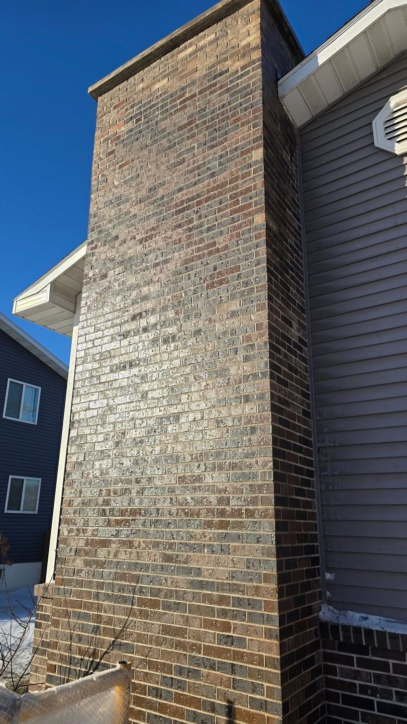 Tall stone chimney on a house exterior with blue sky and neighboring siding.