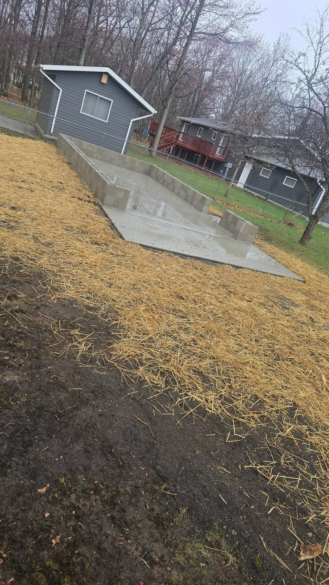 Tilted view of a wet concrete path beside a yellow-leaf-covered hillside, leading to gray buildings in a rainy park