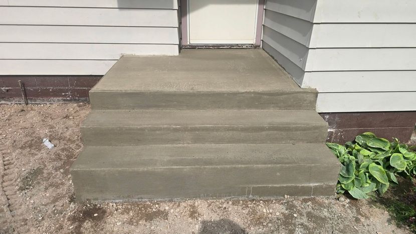Concrete front steps leading to a house entrance, with white siding and green plants on the right.