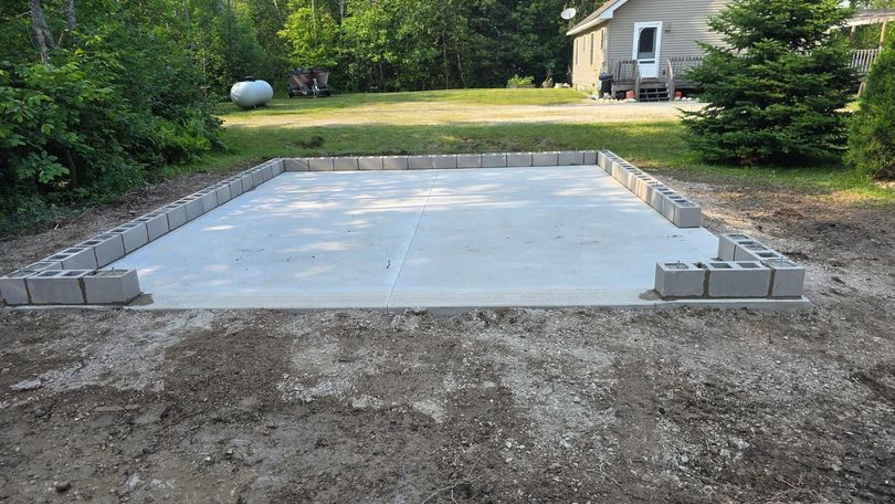Concrete slab foundation framed with cinder blocks in a backyard, near trees and a house