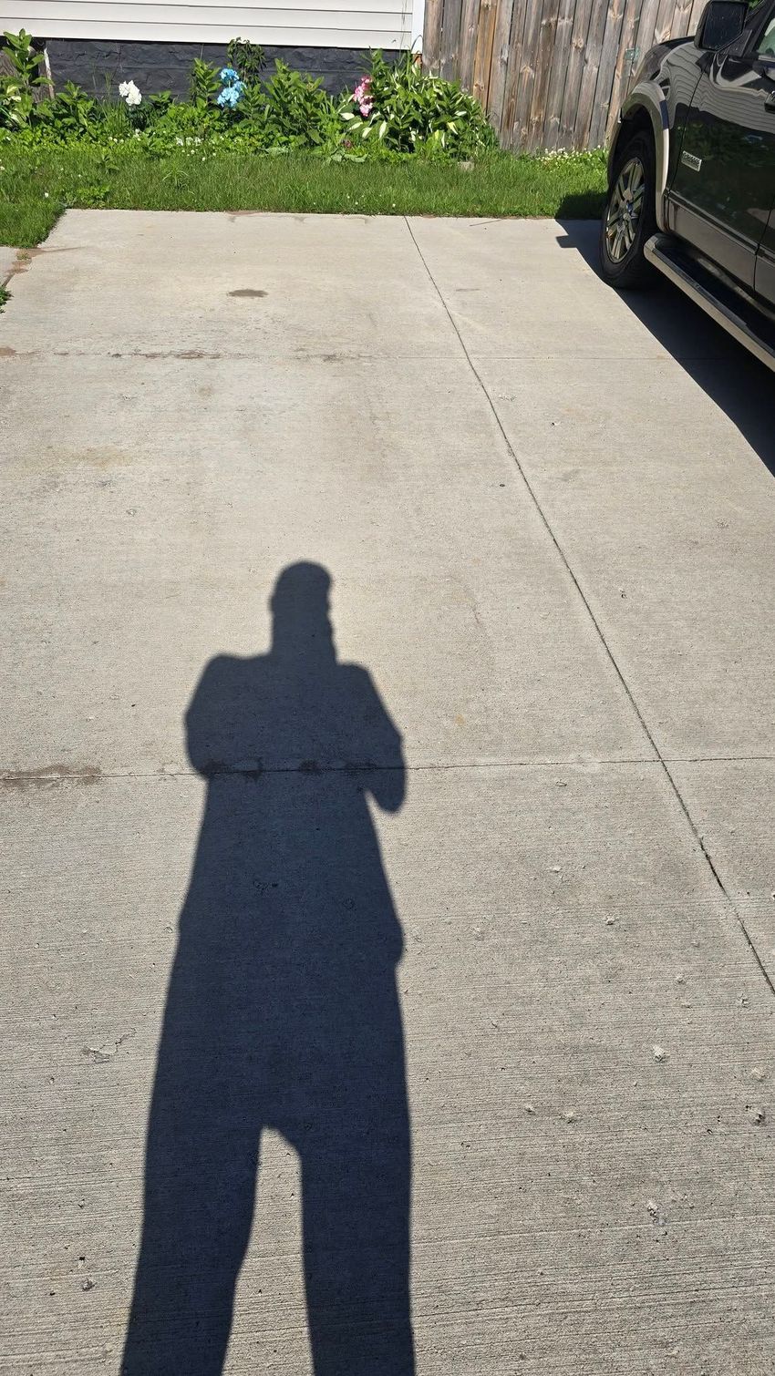 Shadow of a person on a sunlit concrete driveway beside a parked car and a garden bed.