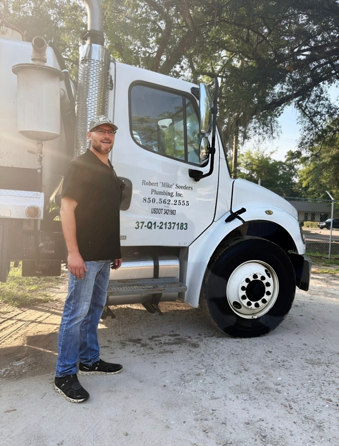 Man standing beside a white utility truck on a gravel lot, with trees in the background.