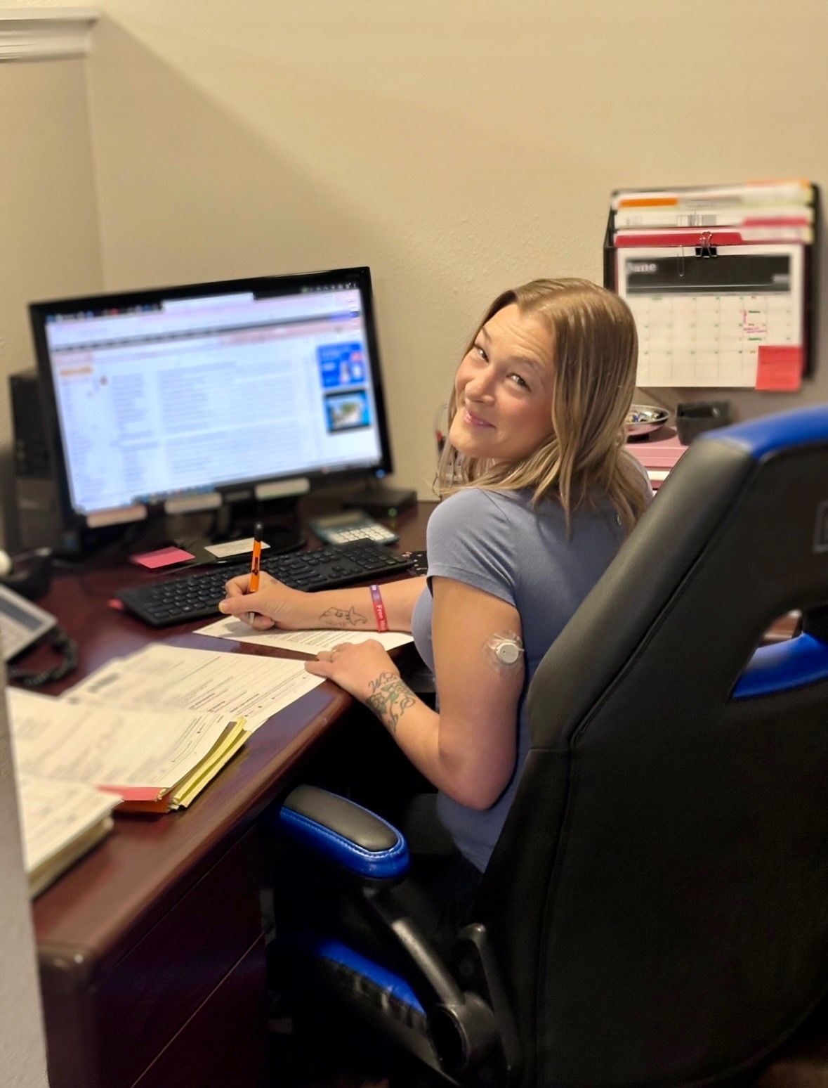 Woman smiling at a desk with computer, papers, and calendar, writing in an office chair.