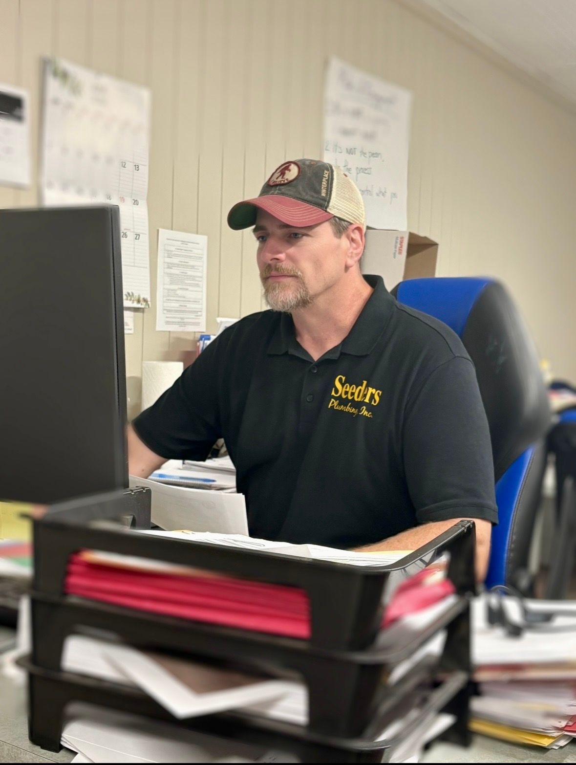 Man in black polo and camo cap working at a desk in an office, with papers and a computer monitor.
