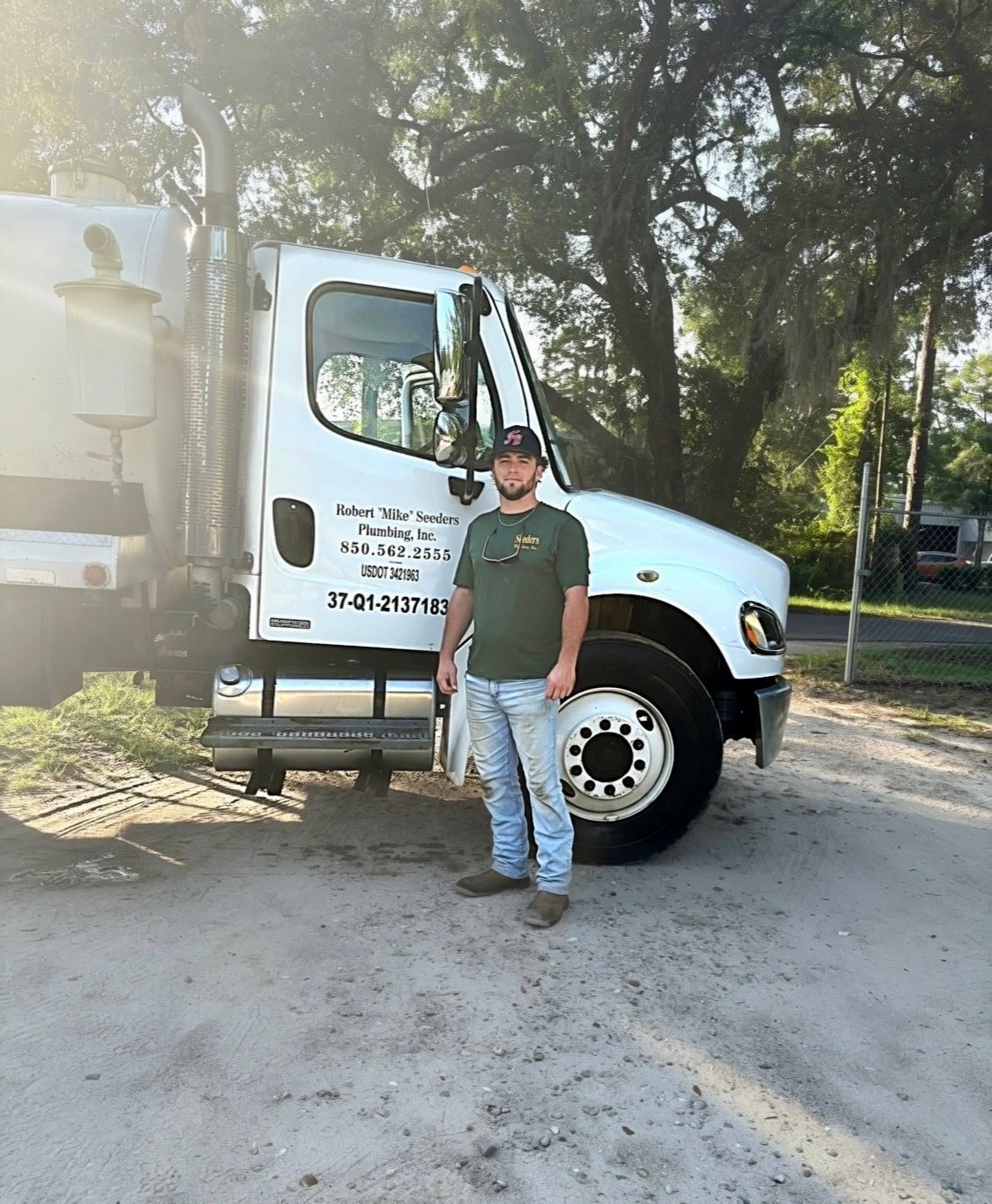 Man standing beside a white delivery truck on a dirt lot under trees