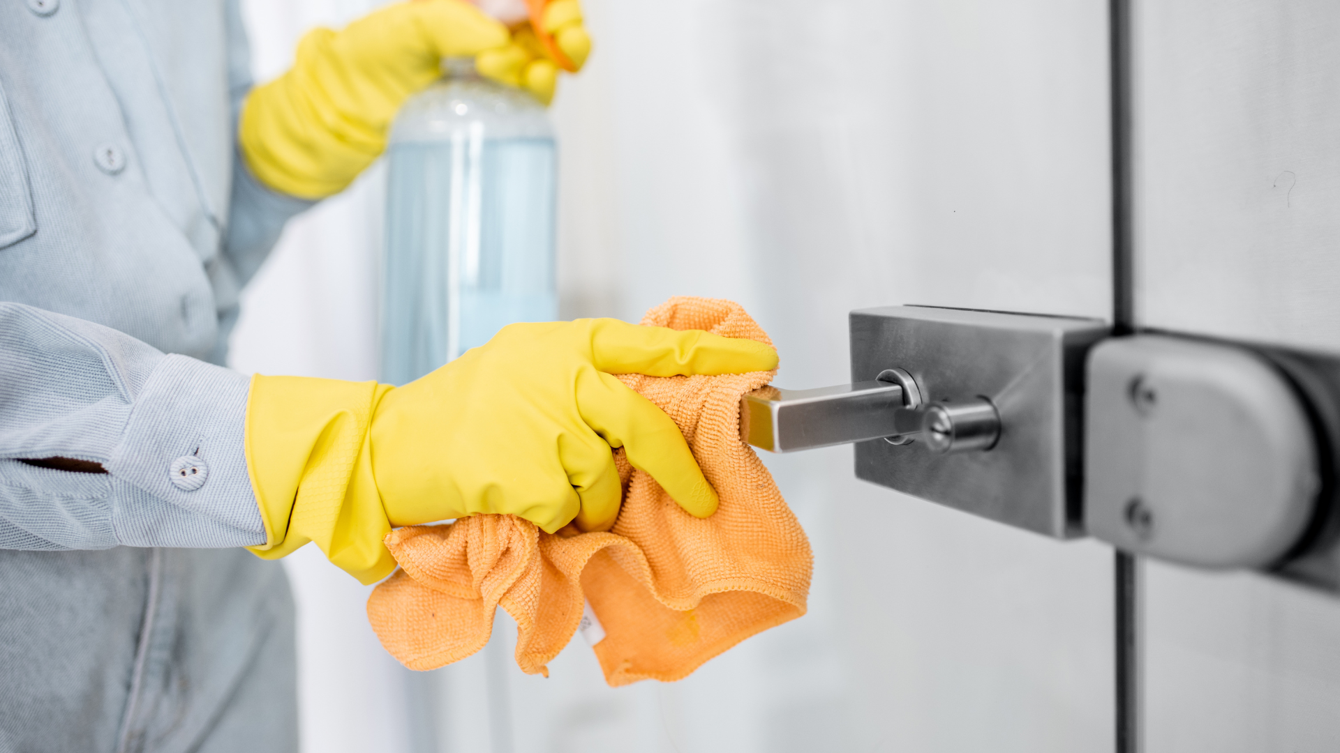 Person wearing yellow gloves spraying and wiping a door handle with an orange cloth.