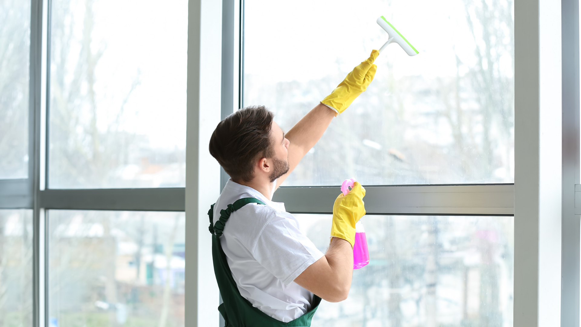 Window cleaner using a squeegee on tall glass panes indoors, wearing yellow gloves and suspenders