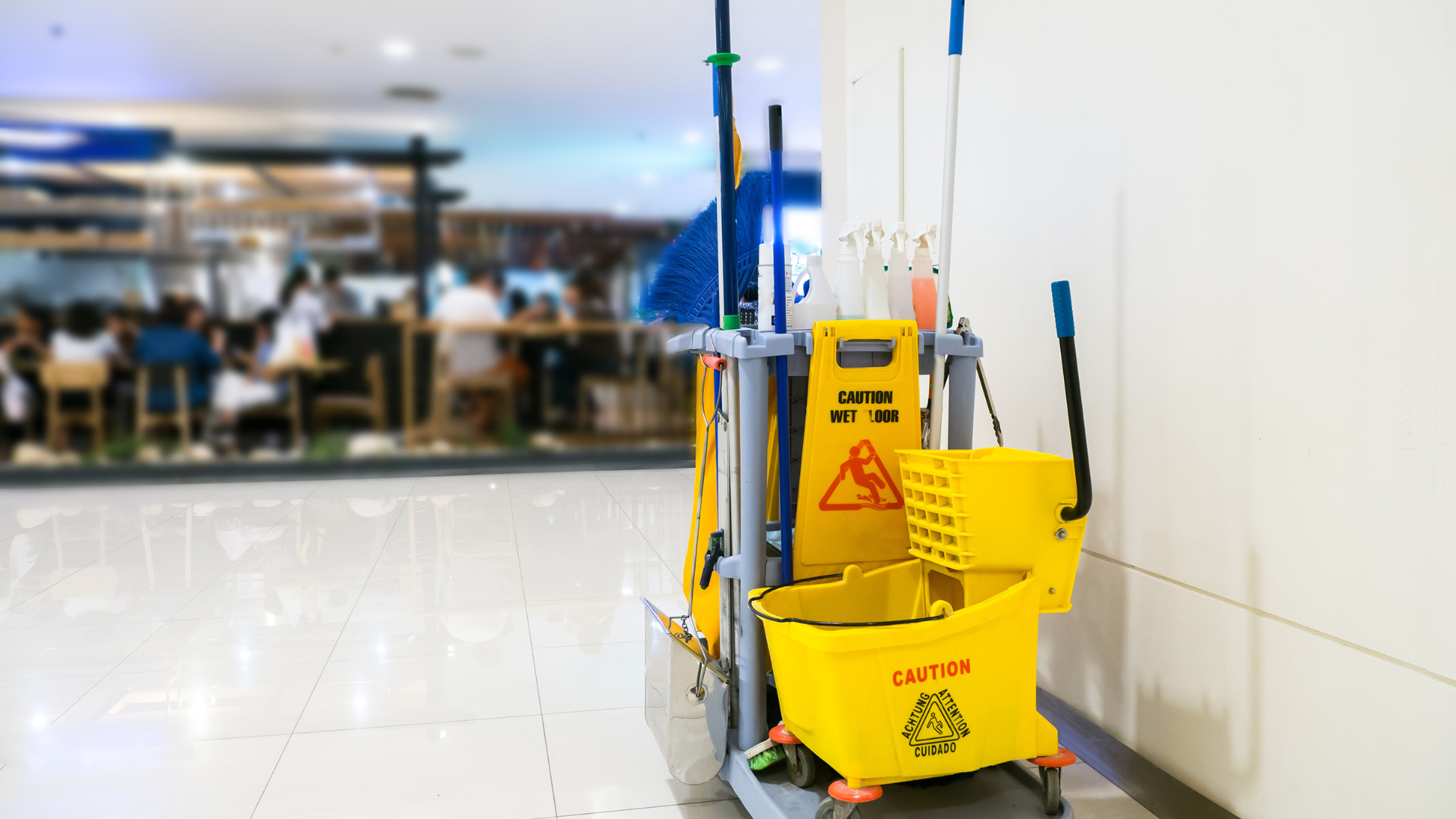 Yellow cleaning cart with caution sign, mops, and buckets, in a brightly lit hallway.