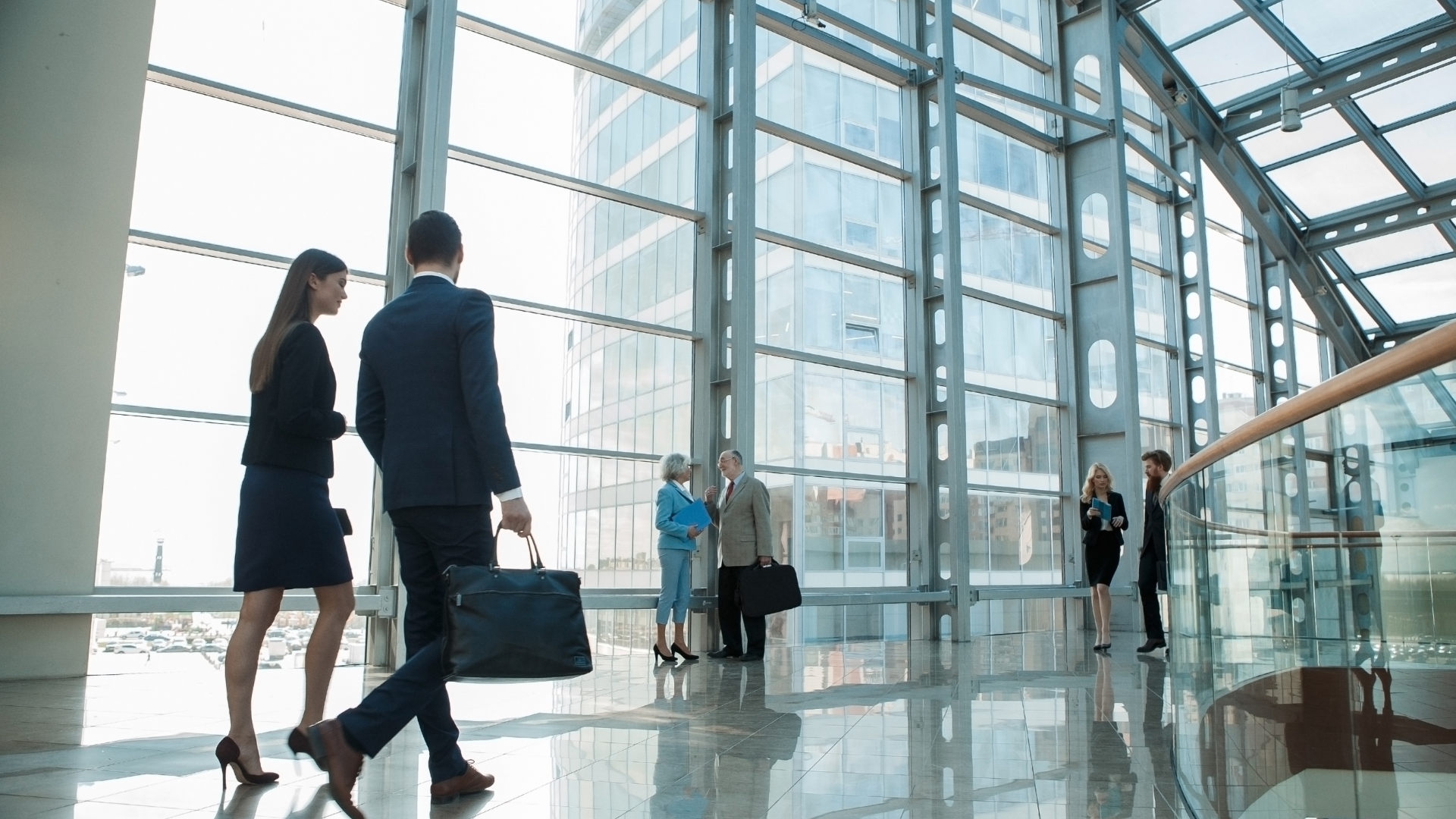 People in business attire walking in a modern, glass-walled office building.