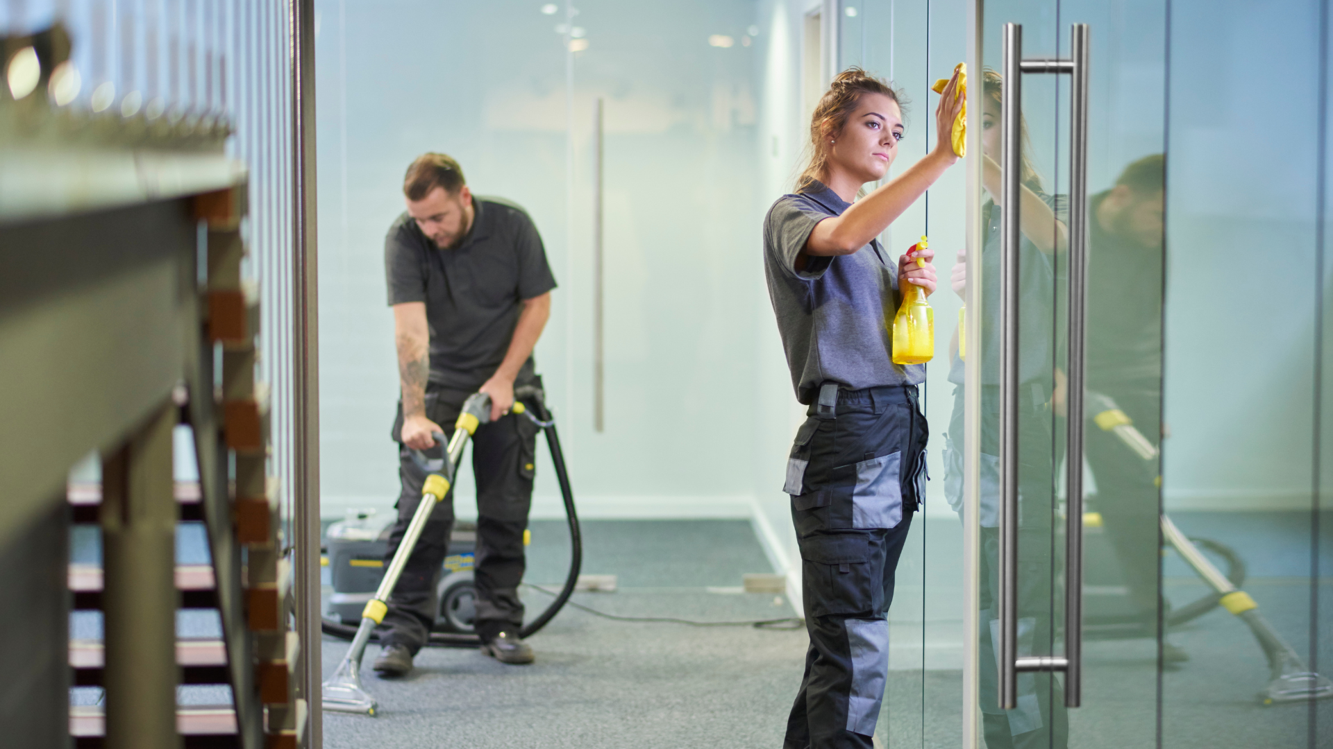 Two workers cleaning a glass wall, one using a spray tool and the other wiping panels.