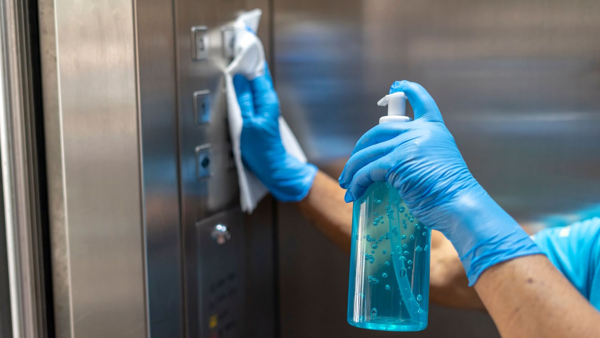 Blue-gloved hands disinfecting an elevator panel with a spray bottle and cloth