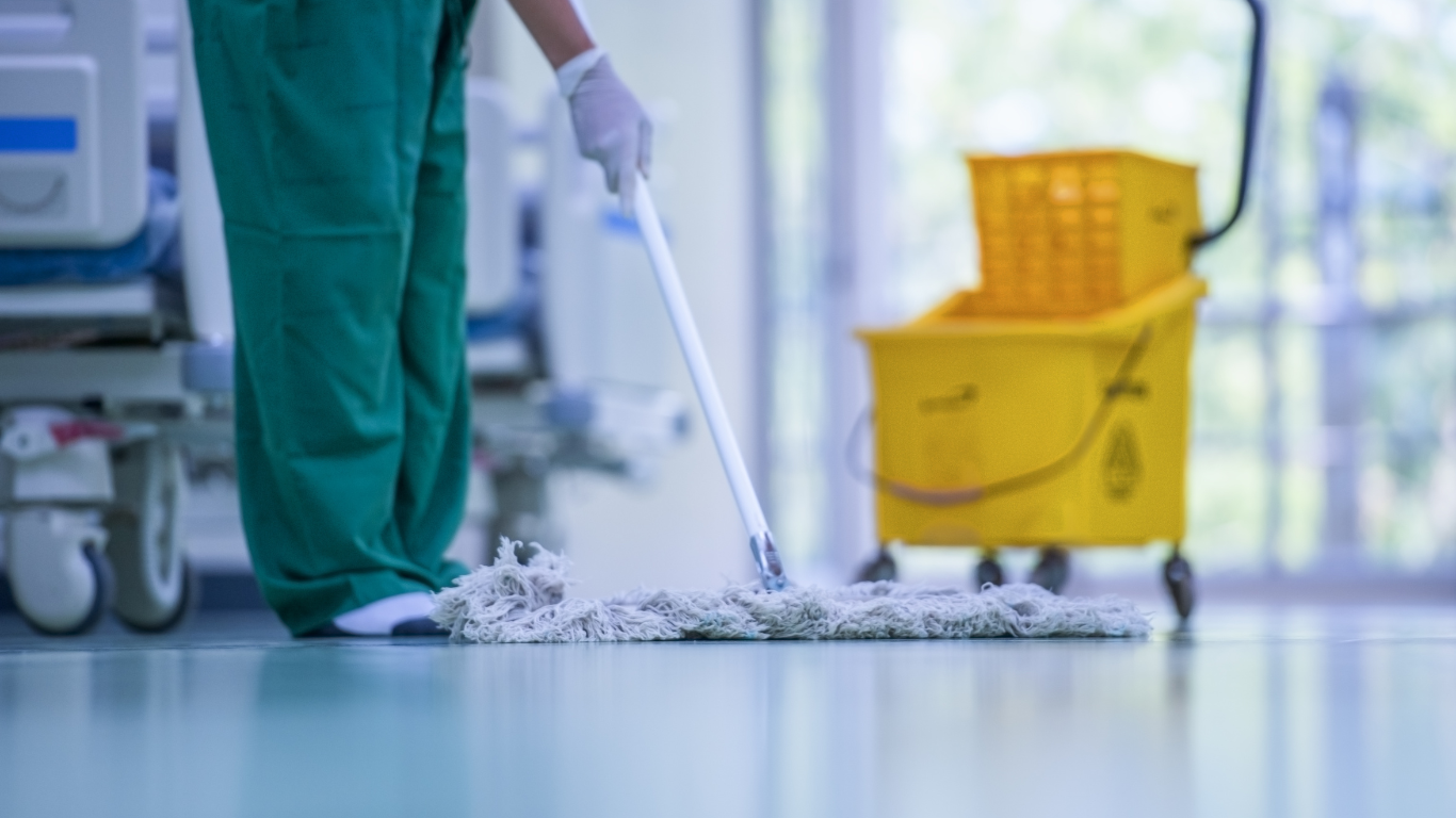 Person in green scrubs mops a hospital floor near a yellow cleaning bucket and hospital bed.
