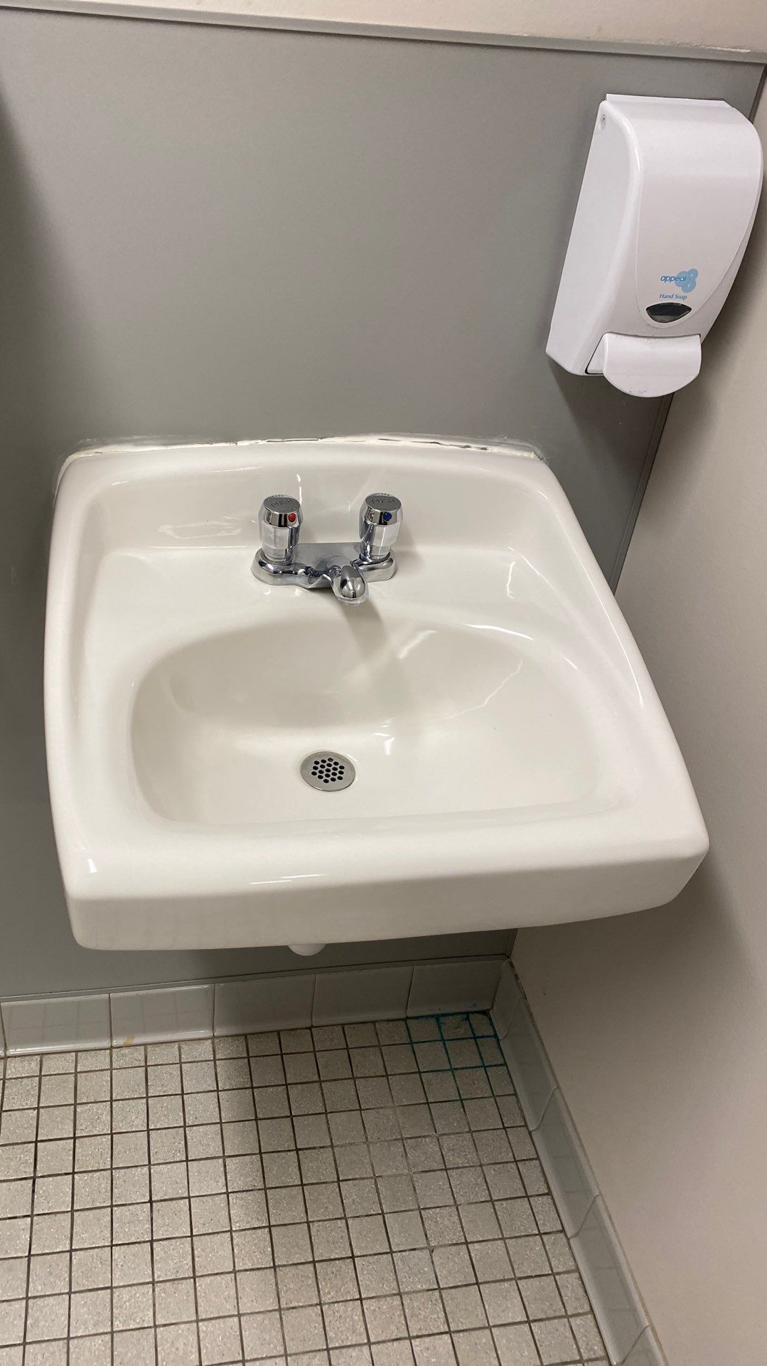 White sink with chrome faucet and soap dispenser on gray wall. Small tiled floor.