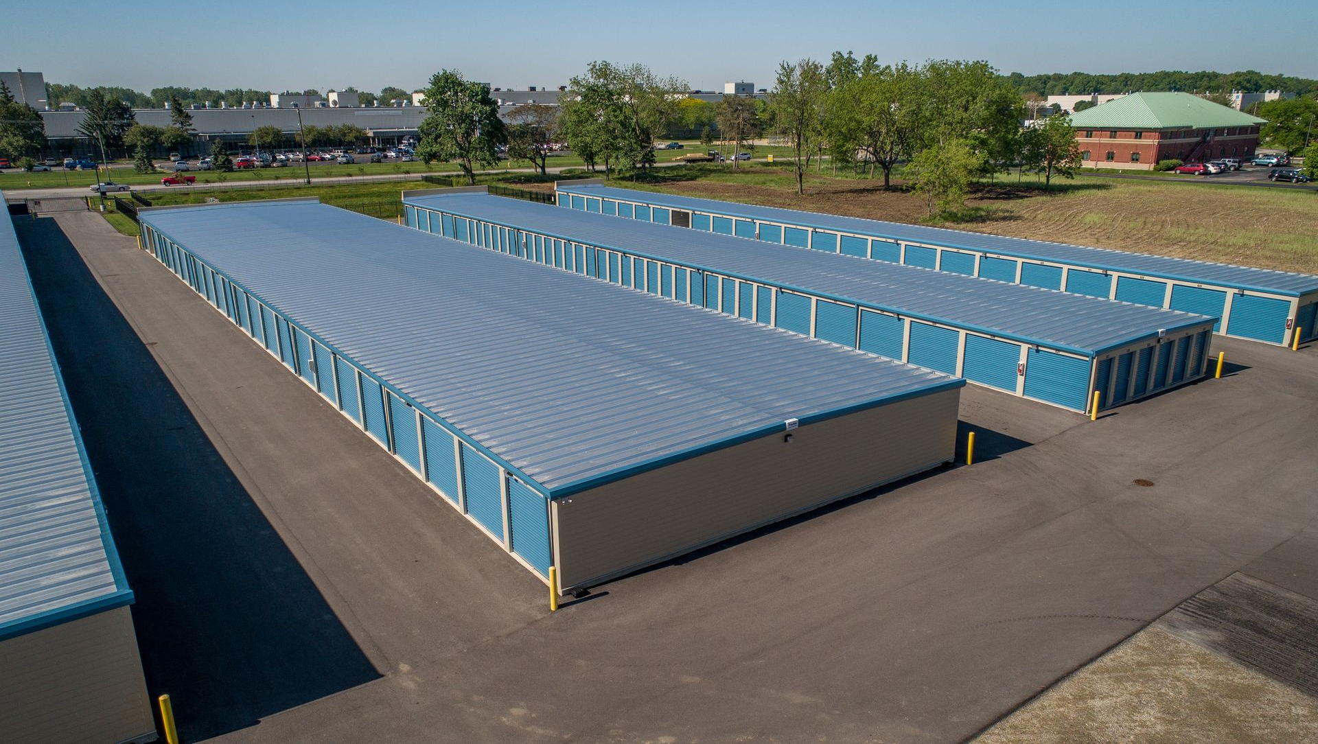 Aerial view of rows of blue storage units in a paved lot beside a grassy area and trees.