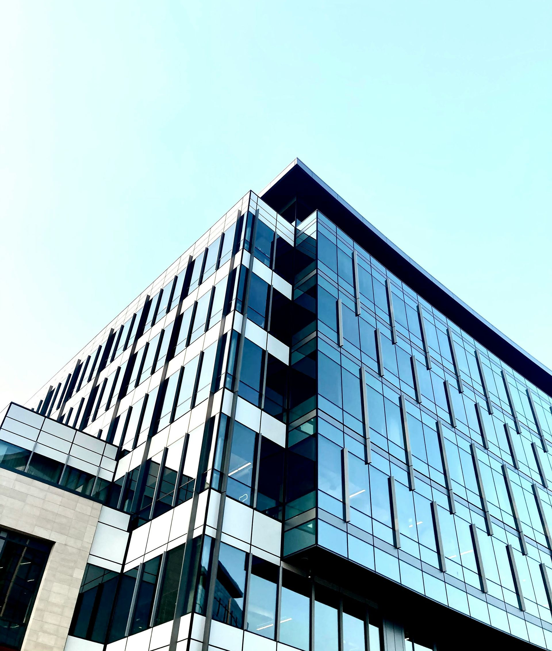 Modern glass and steel building, angled upward with a pale blue sky in the background.