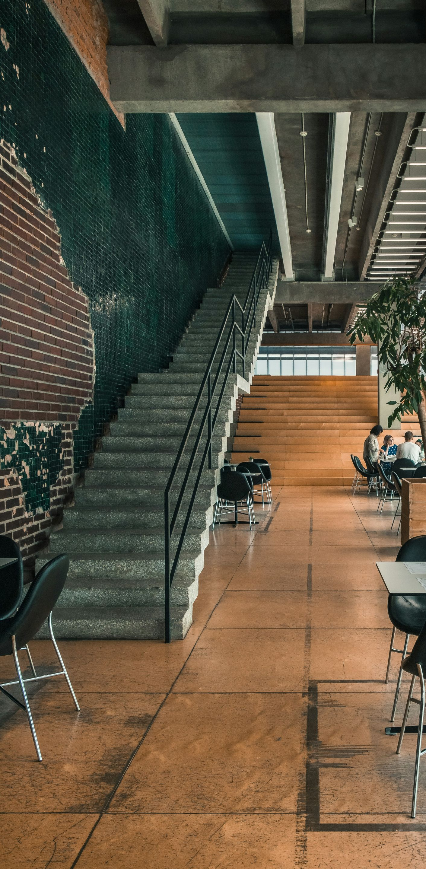 Modern lounge with staircase, wooden floor, black chairs, long tables, and indoor plants.