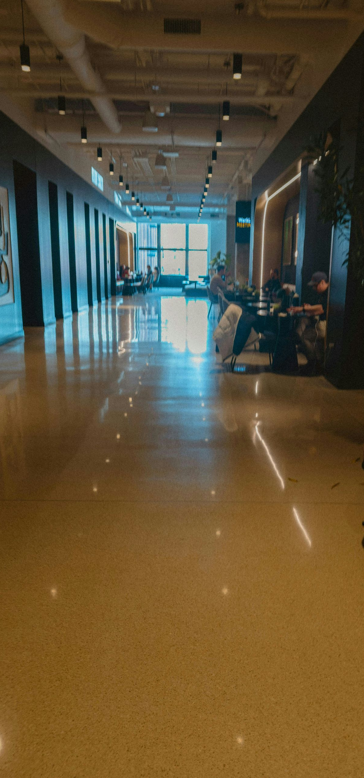 Dim hallway with glossy floors, wall art, and chairs along the sides, lit by bright windows at the far end.